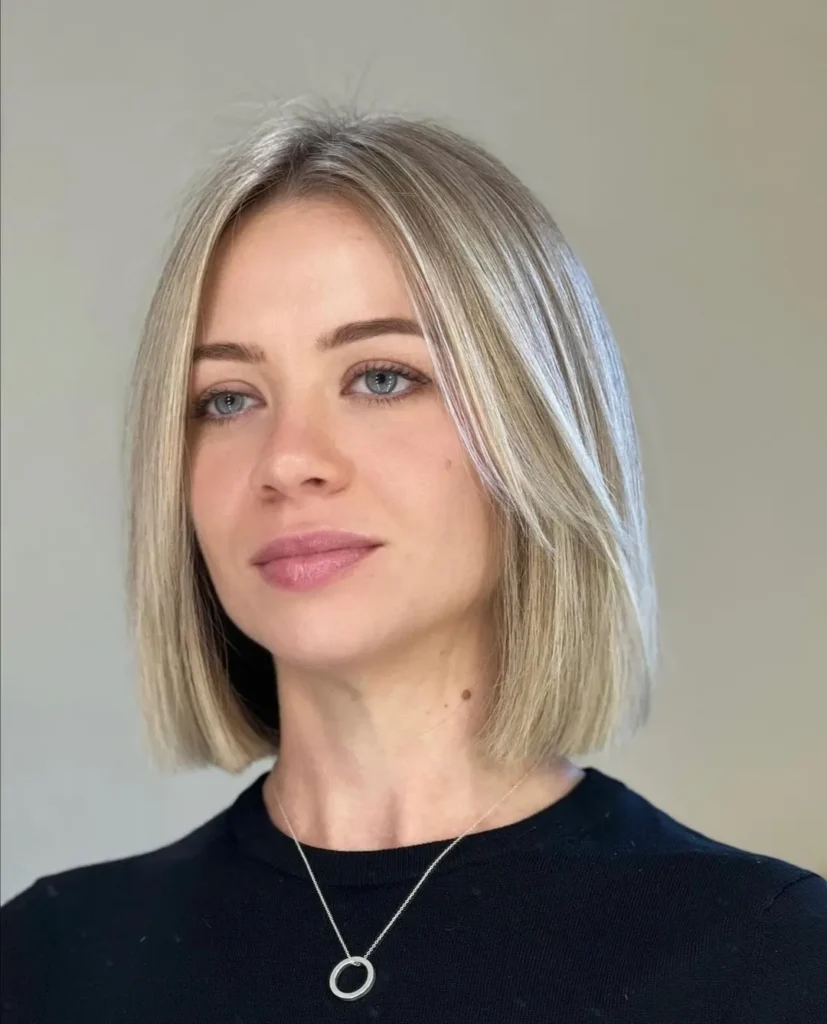 A woman with a sleek icy blonde middle-part lob wears a black top and a circle pendant necklace.