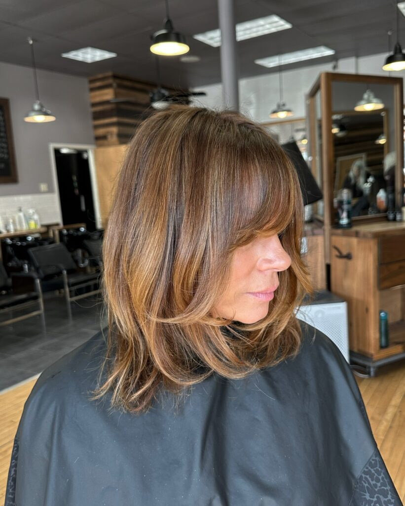 A woman with medium-length brown hair featuring caramel balayage, curtain bangs, and soft layered ends styled at a hair salon.