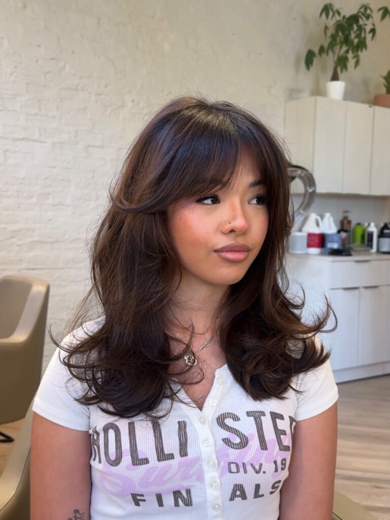A woman with medium length brown hair styled into voluminous curls with wispy curtain bangs.