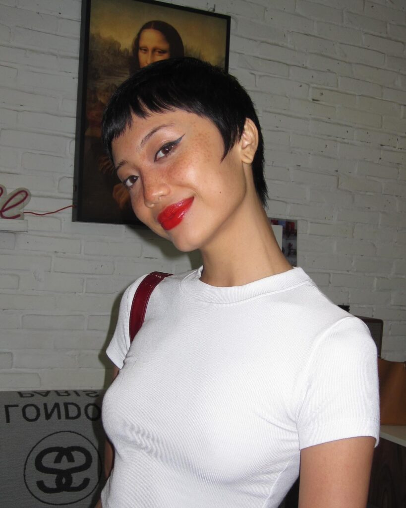 A woman with a sleek black pixie cut and blunt bangs smiles over her shoulder wearing red lipstick.