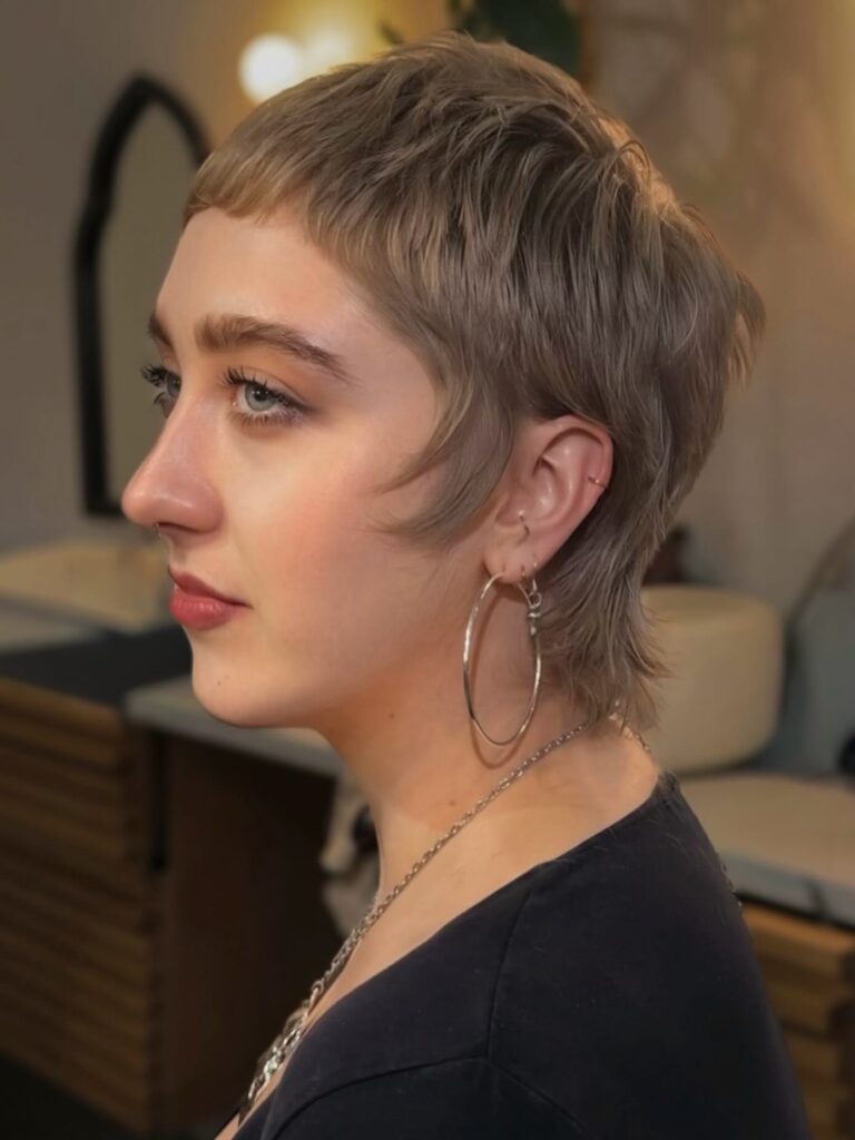 A woman with a sandy blonde mullet pixie cut and silver hoop earrings is shown in profile.