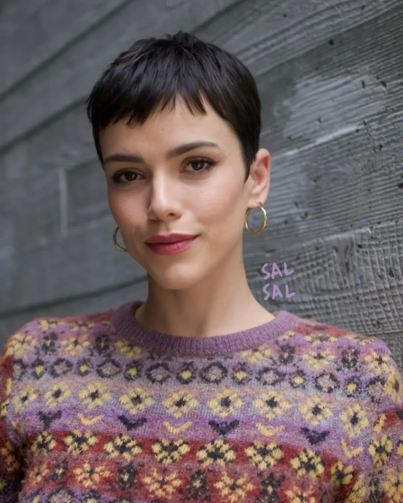 A woman with a short black pixie cut and tiny blunt bangs wears gold hoop earrings and a patterned sweater.
