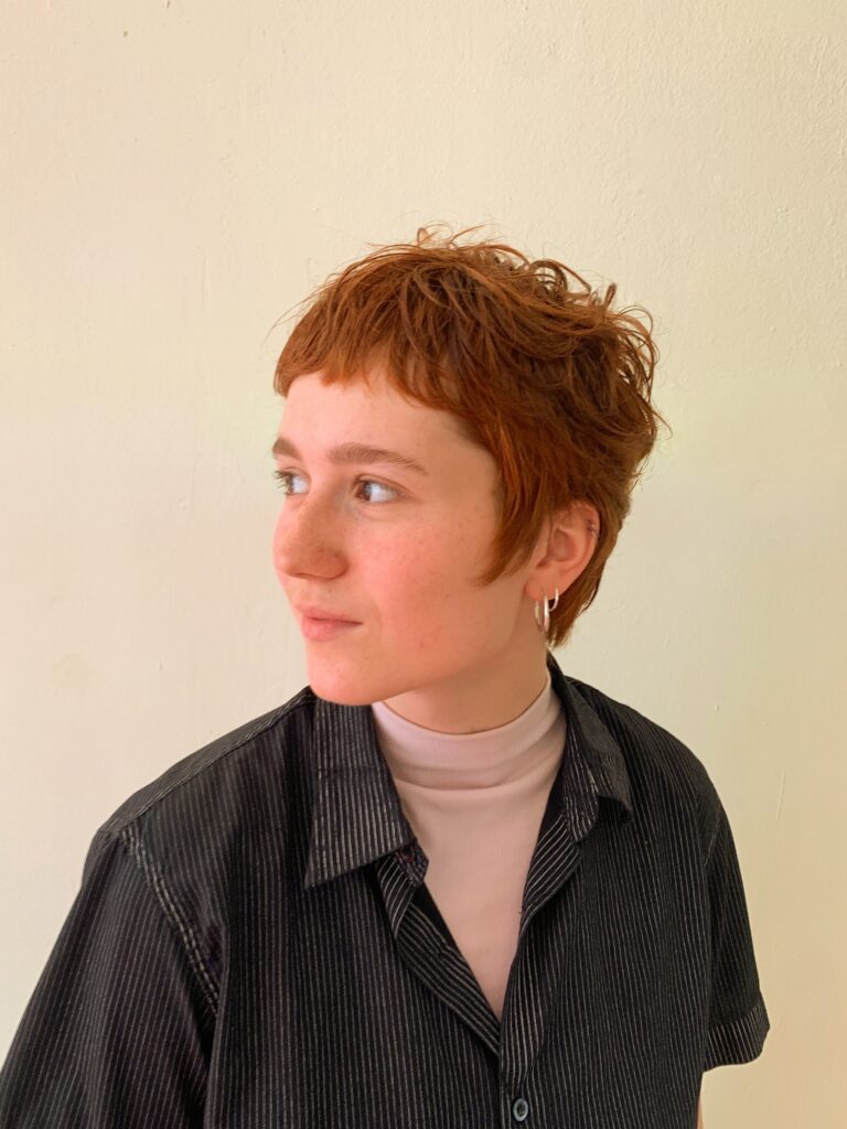 A woman with a tousled copper pixie cut and curtain fringe looks to the side wearing small hoop earrings.