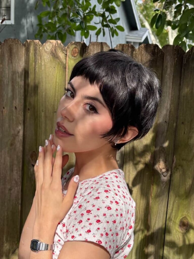 A woman with a sleek black pixie cut featuring full blunt bangs poses outdoors near a wooden fence.