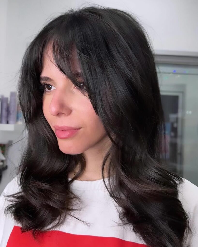 A woman with a glossy black blowout featuring deep side-swept curtain bangs and voluminous flipped ends.
