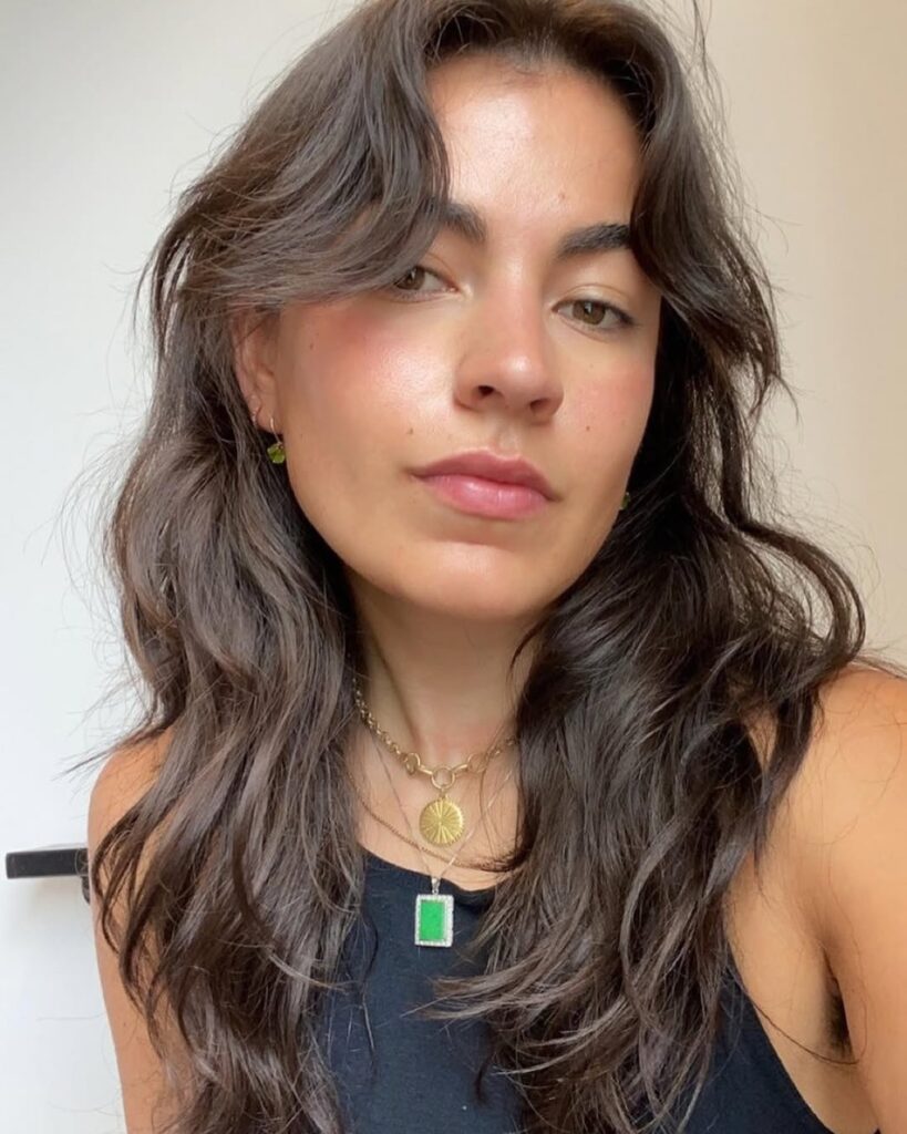 A woman with long brunette waves and wispy curtain bangs styled in a relaxed, undone texture.