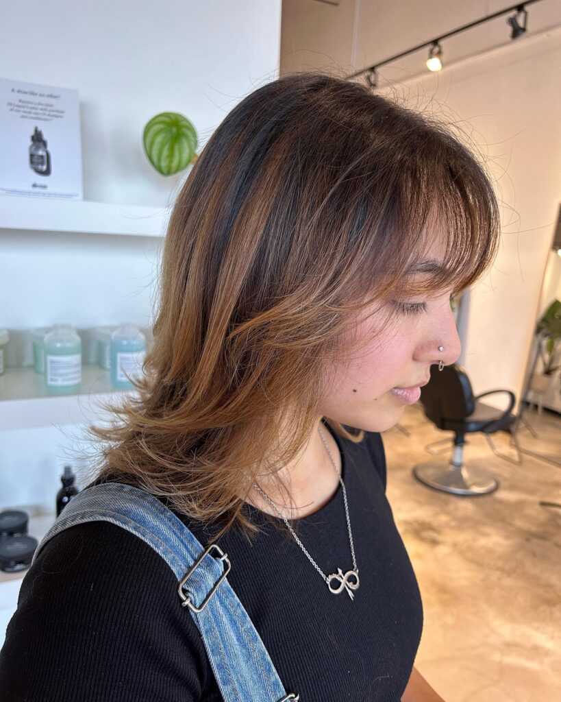 A woman with a shoulder-length brunette shag featuring caramel balayage and curtain bangs.