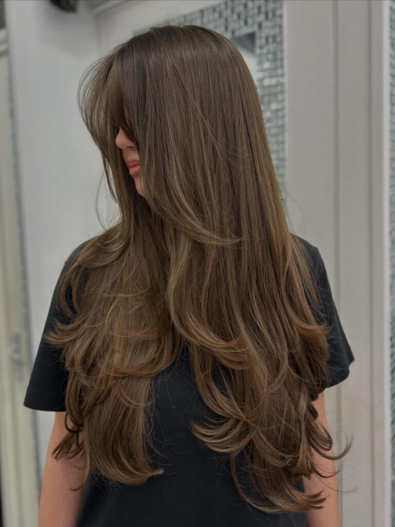 Long ash brown hair styled in chunky flipped-out butterfly layers with wispy face-framing bangs.