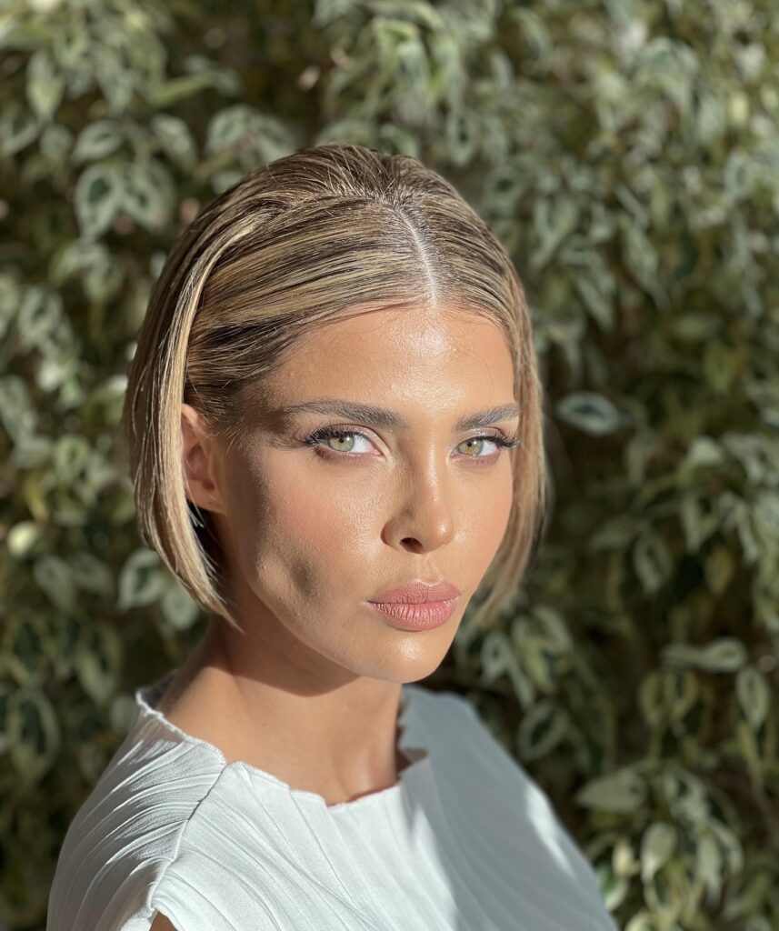 A close-up portrait of a woman with sleek, center-parted blonde hair, green eyes, and natural makeup against a leafy outdoor background.