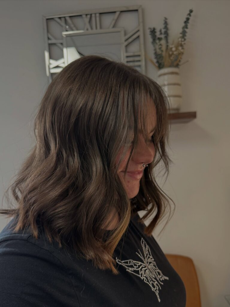 A woman with shoulder-length wavy brunette hair featuring caramel highlights, soft layers, and curtain bangs framing her face.
