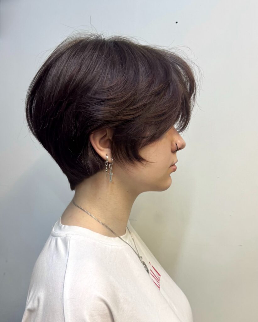 A woman with a rounded brunette layered bob and curtain bangs wears dangly earrings and a white shirt.