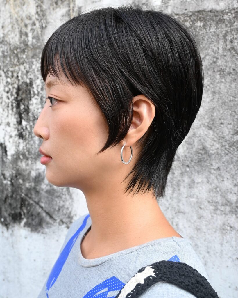 A woman with a sleek black long pixie cut and blunt fringe wears silver hoop earrings against a textured wall.