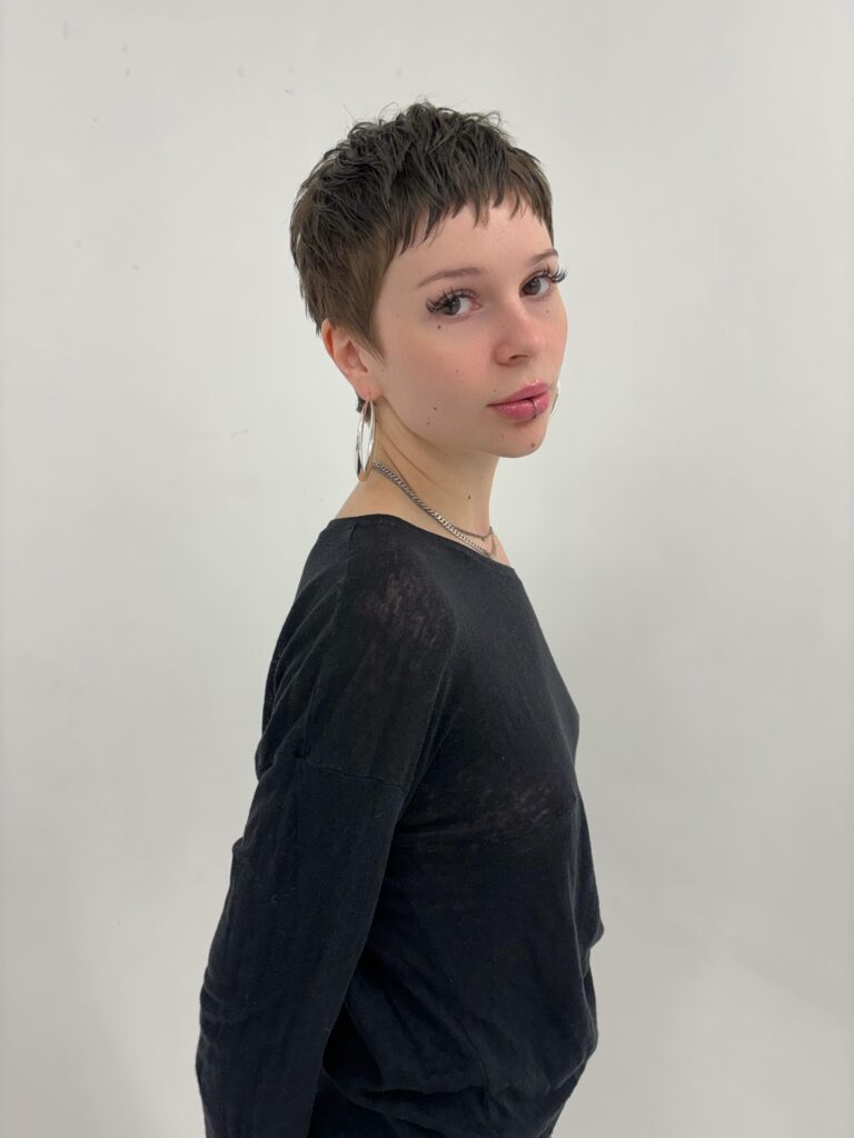 A woman with a short brunette textured pixie cut and blunt fringe poses in profile wearing hoop earrings and a black top.