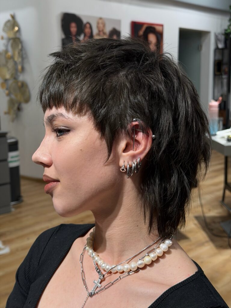 A woman showing her side profile with a dark brown choppy mullet haircut featuring micro bangs and layered textured crown.