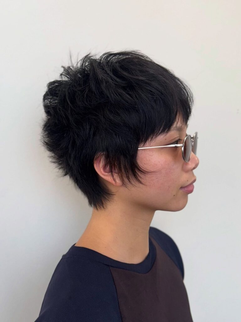 A woman with a short black voluminous textured crop wears round sunglasses and a dark shirt.