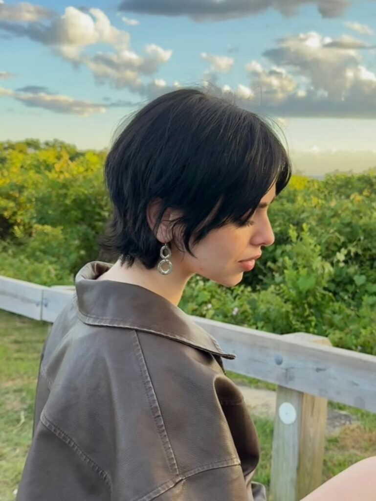 A woman outdoors wearing a leather jacket featuring a soft black bixie haircut with curtain bangs and feathered layers.