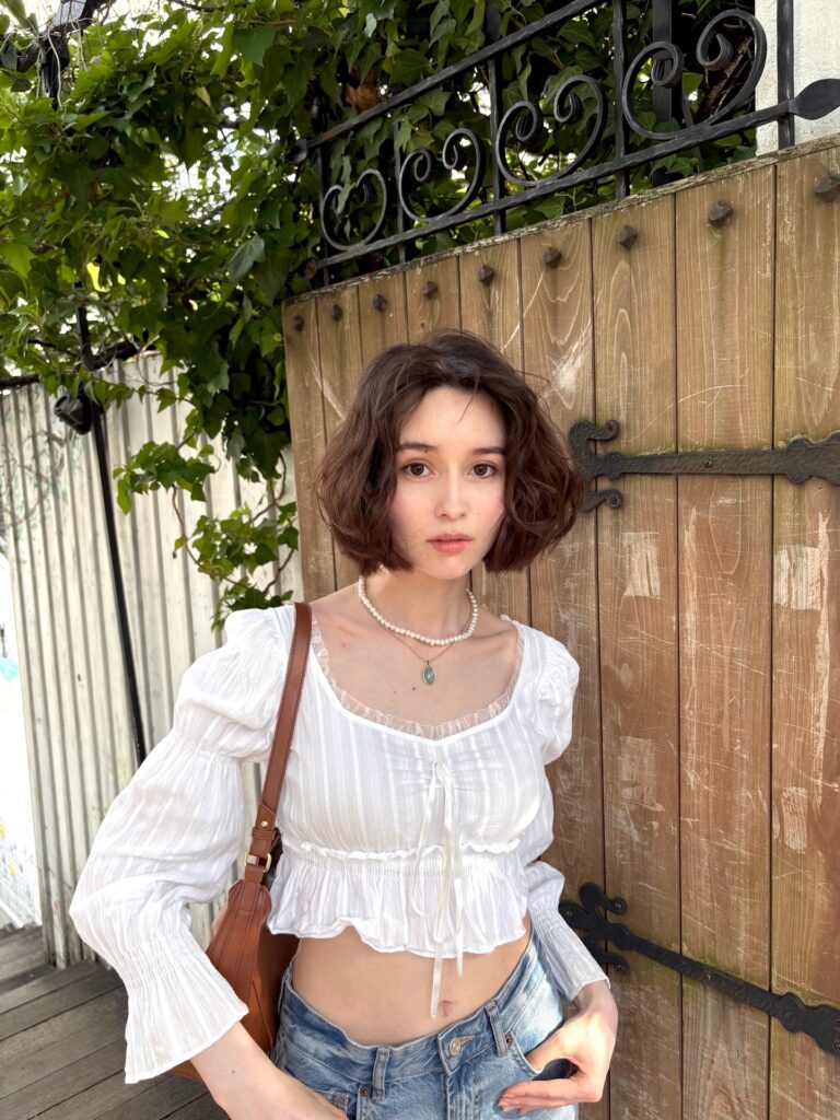 A woman with a chin length curly brown bob and pearl necklace stands in front of a wooden garden gate.