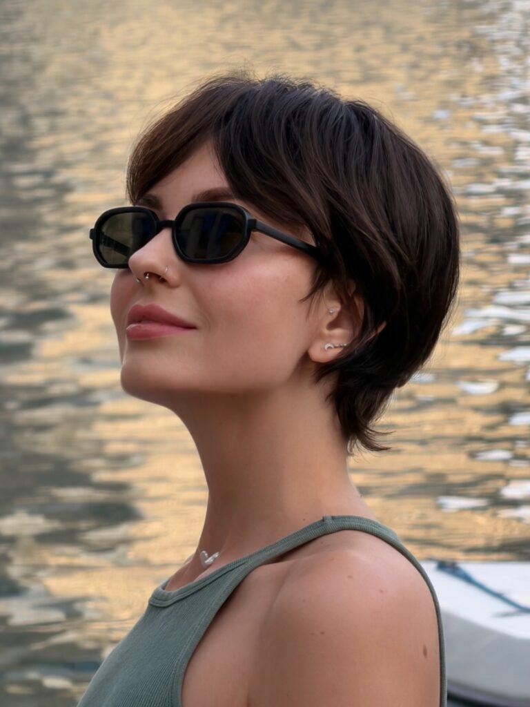 A woman with a dark brown wavy pixie cut and long side-swept bangs wears sunglasses while looking off to the side near the water.