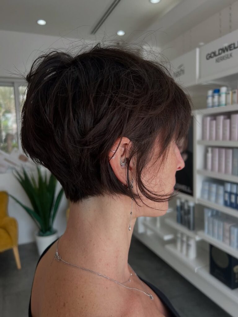 A woman with a dark brown tousled jaw length bob and delicate ear piercings looks off to the side in a salon.