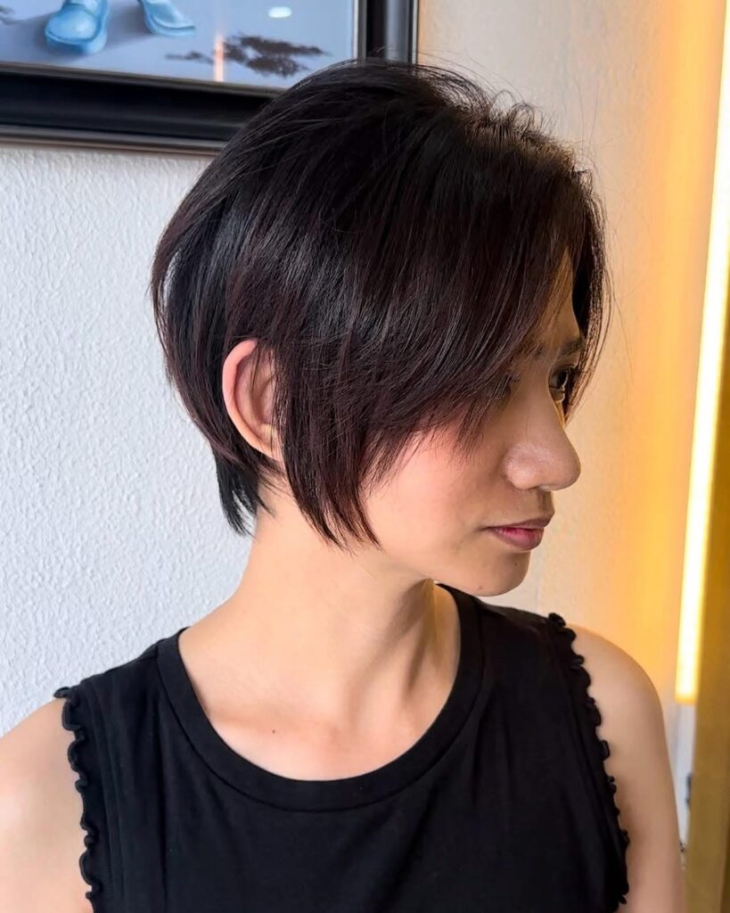 A woman with a long dark brown pixie cut and deep side swept fringe looks off to the side in profile.