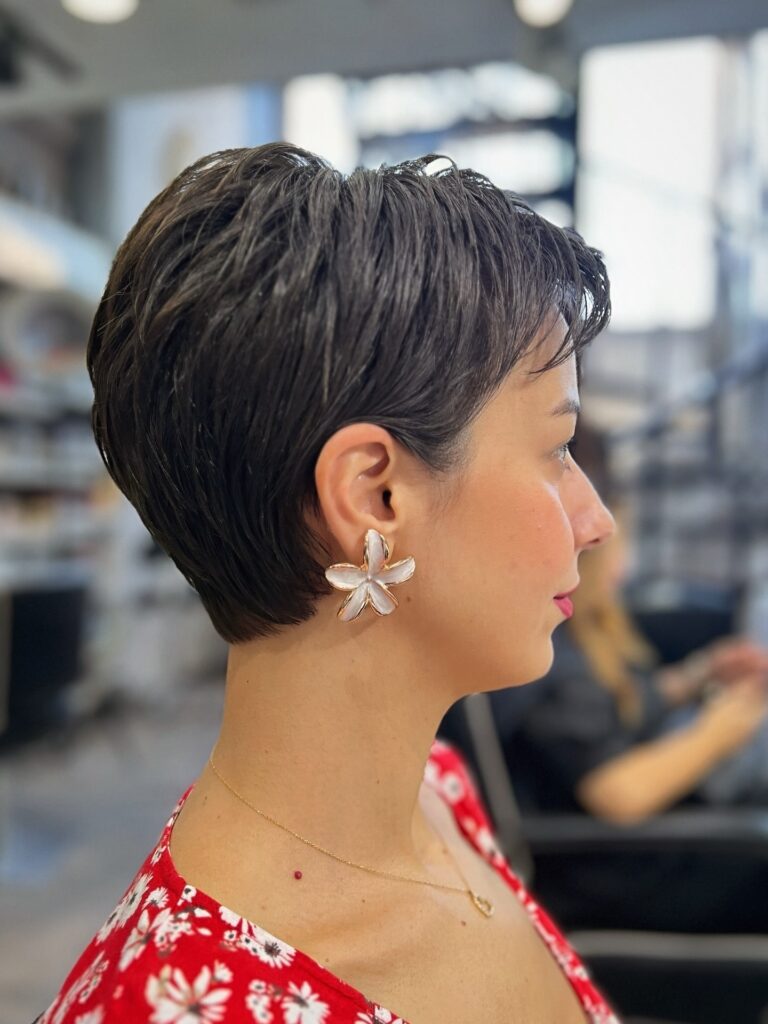 A woman with a sleek dark brown cropped pixie and flower earring is shown in profile at a salon.