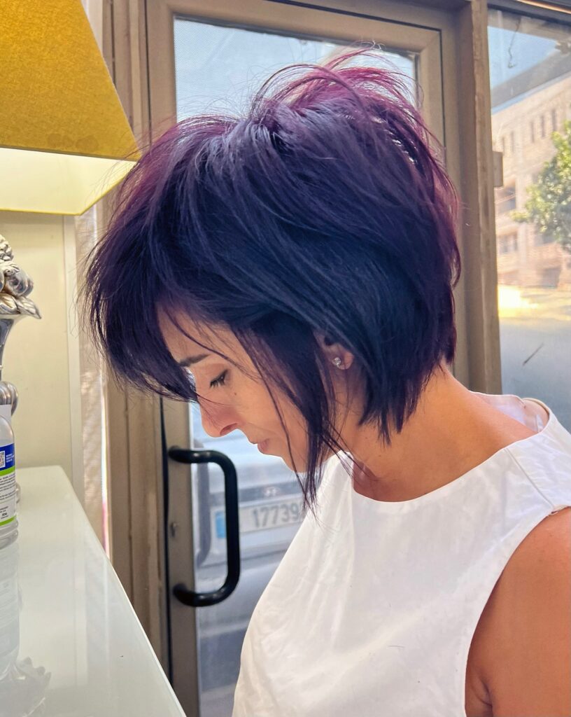 A woman with a choppy purple and black shag haircut looks down while wearing a white top indoors.