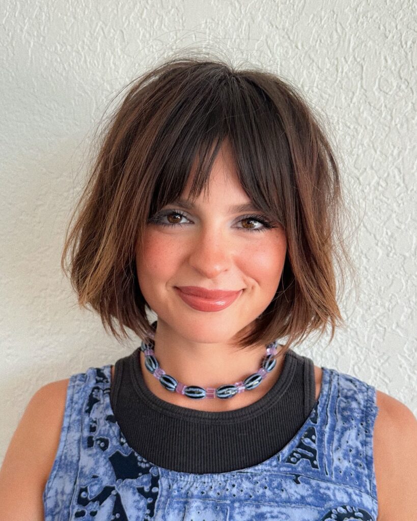 A woman with a chin length brunette bob and wispy see through bangs smiles while wearing a beaded necklace.