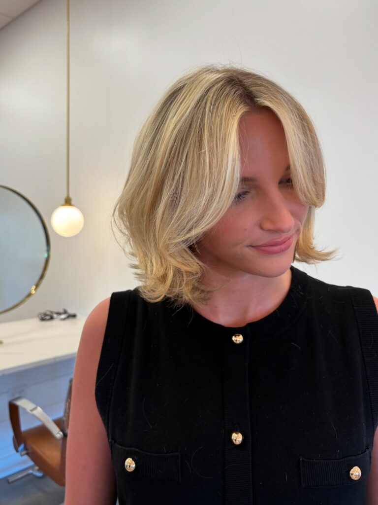 A woman with a wavy buttery blonde bob and dimensional highlights looks down and smiles softly.