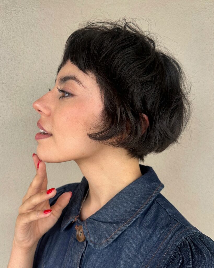  A woman with a rounded brunette bob and short micro bangs is shown in profile touching her chin.