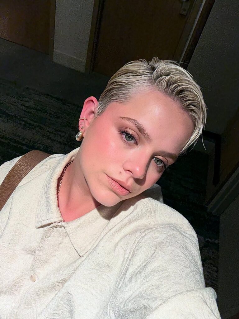 A woman with a slicked back platinum blonde pixie cut and ear piercings looks at the camera in dim lighting.