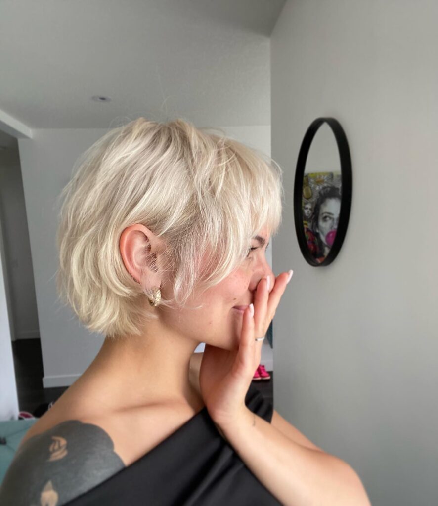 A woman with a short choppy platinum blonde bob and curtain bangs smiles while touching her face at home.