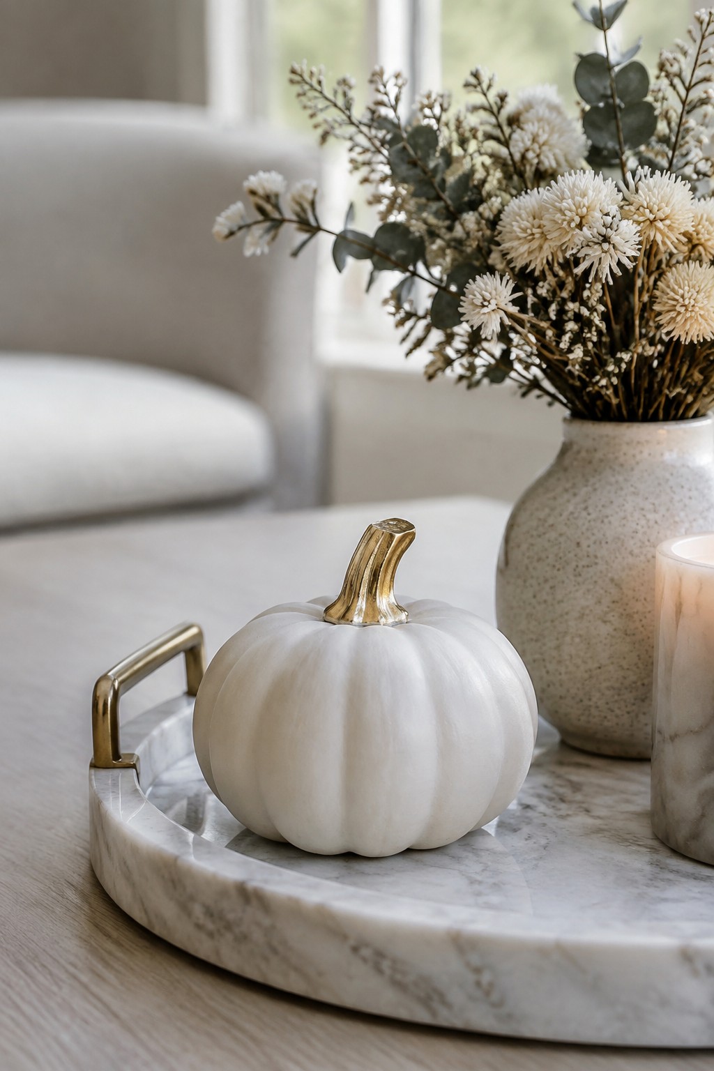 A small white pumpkin with a gold stem sits on a marble tray next to a speckled vase and candle.