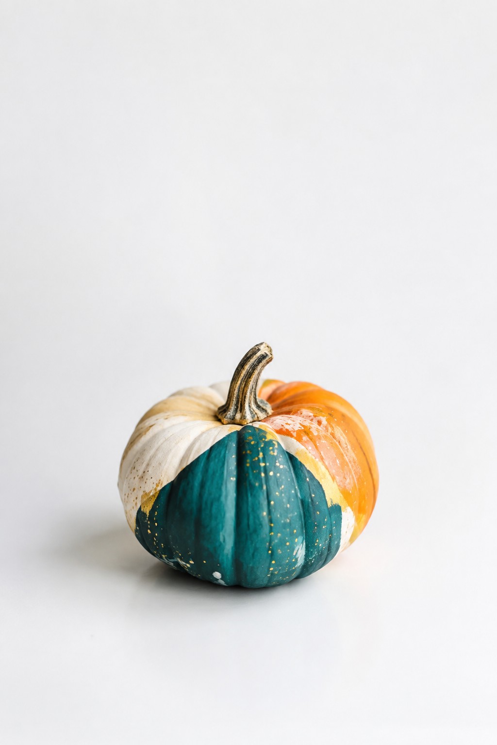 Small pumpkin painted in teal, white, and orange sections with gold splatters on a plain white background.