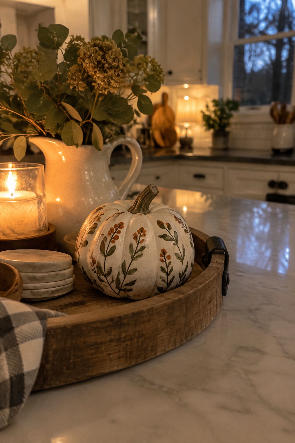 A white mini pumpkin painted with orange and green botanical patterns rests in a wooden tray on a marble kitchen counter.