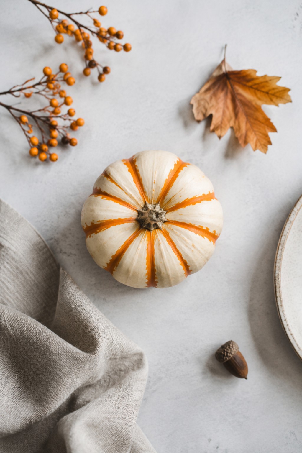A small white pumpkin with orange painted stripes rests on a light surface alongside berry branches, a dried leaf, and an acorn.