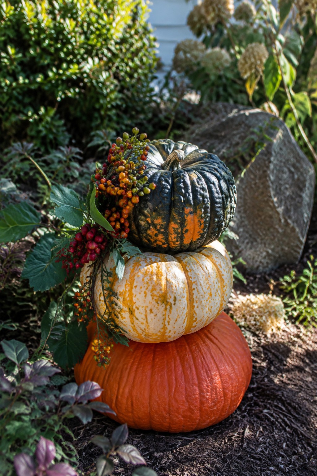 Three pumpkins stacked vertically outdoors with an orange base, white middle, and dark green top, accented with red and orange berries in a garden setting.