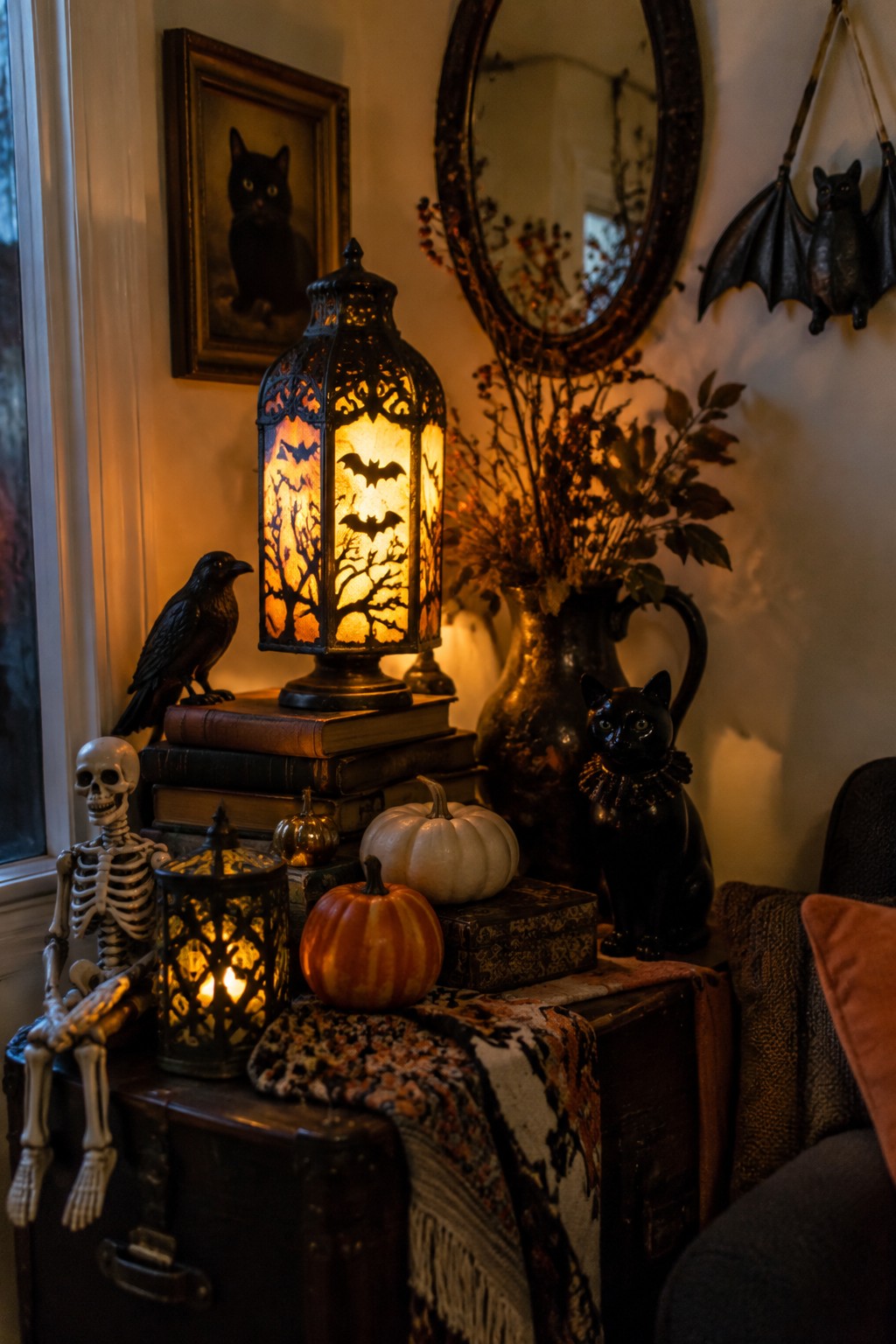 A glowing black metal lantern with bat and tree cutouts sits on stacked books atop a trunk, next to a skeleton, black cat figurine, crow, and small pumpkins.