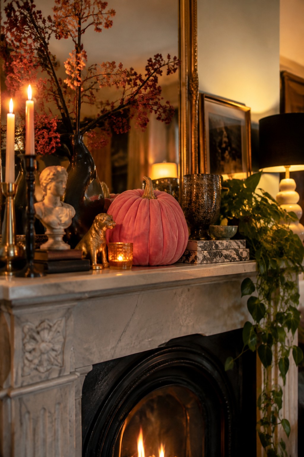 A large pink velvet pumpkin sits centered on a white marble fireplace mantel with lit candles, a small golden dog figurine, and dark branches in a black vase.