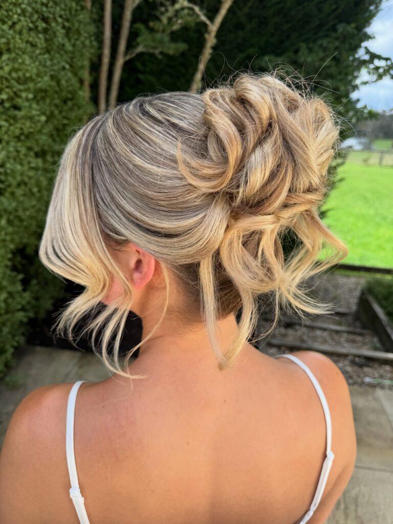 A woman with blonde curly hair styled in a textured updo with wispy face-framing pieces.