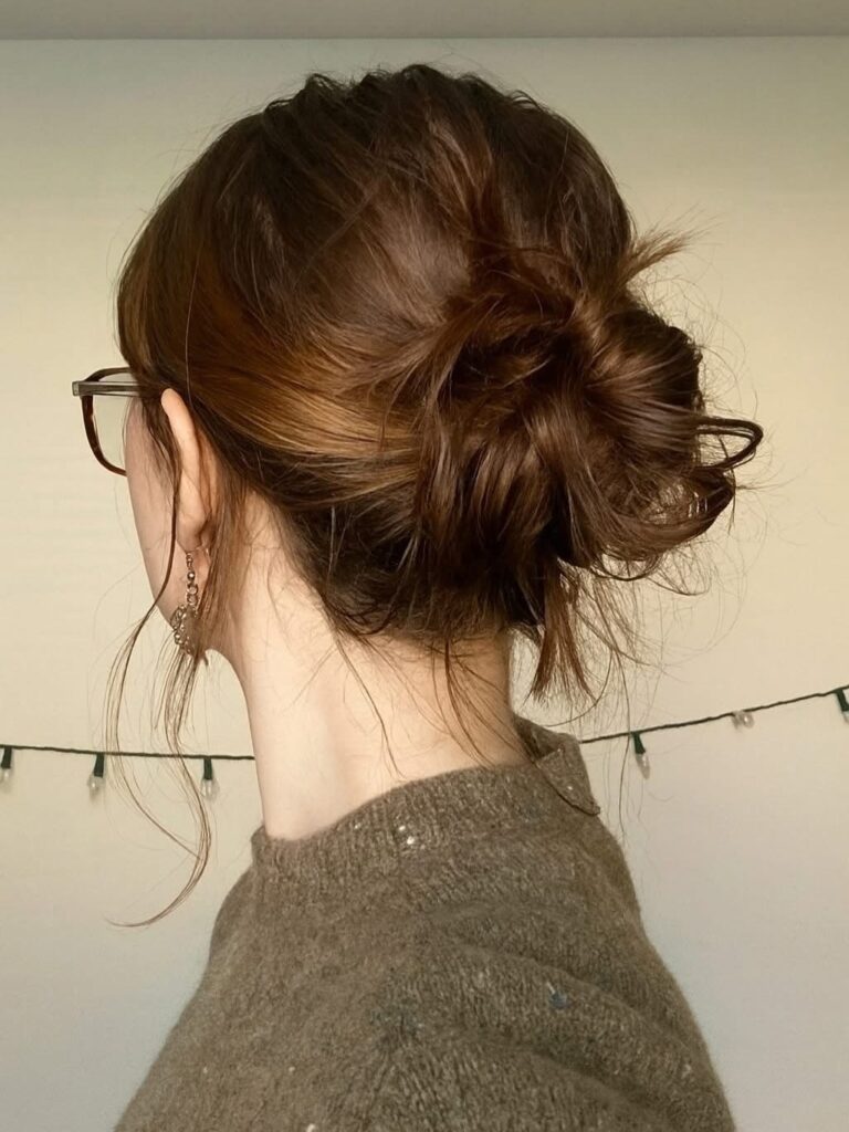 A woman with auburn brown hair styled in a low messy bun with loose strands framing her neck and ears.