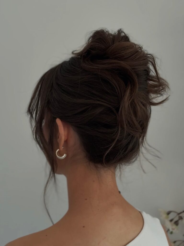 A woman with dark brown hair styled in a voluminous messy bun with wispy layers framing her neck and face.