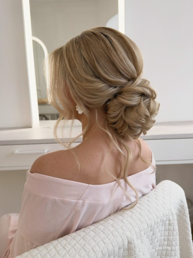A woman with blonde hair styled in a textured rose shaped updo with soft waves framing the face.