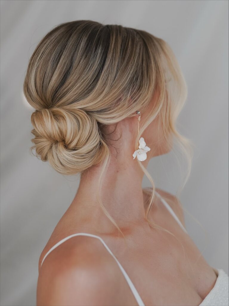 A woman with blonde balayage hair styled in a sleek low twisted bun with loose face-framing strands.