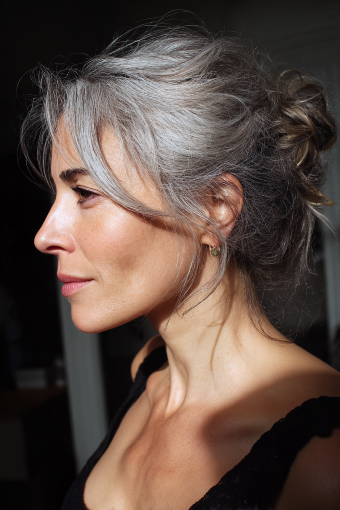 Side profile of a woman with natural silver-gray hair styled in a textured, messy low bun with soft, face-framing curtain bangs.