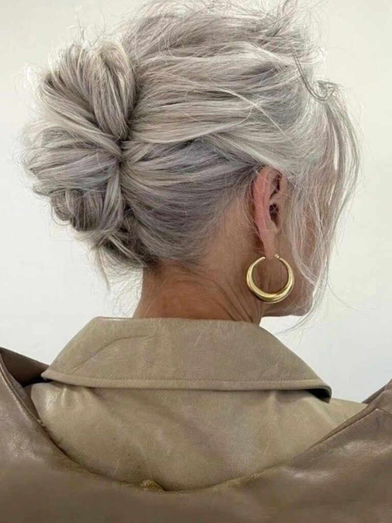 A woman with gray and silver hair wears a loose textured bun with face-framing pieces and gold hoop earrings.