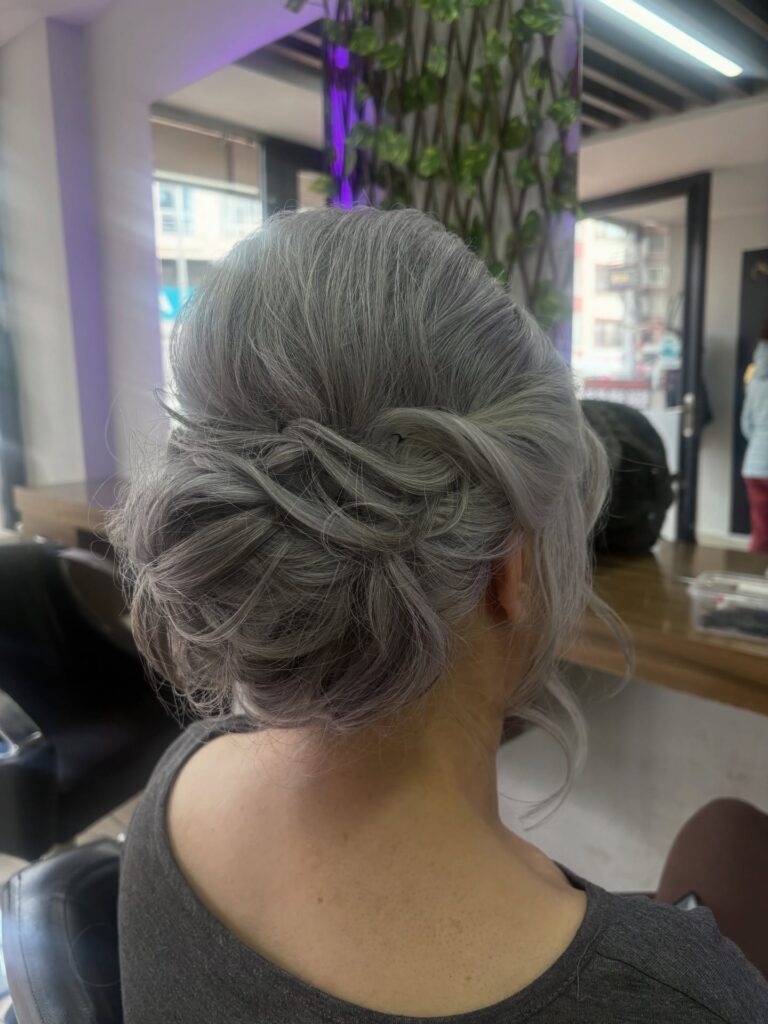  A woman with silvery gray hair wears a soft undone low chignon with wispy face-framing layers.