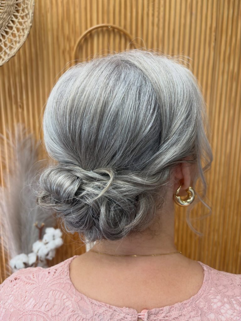 A woman with silvery gray hair wears a loose knotted low bun paired with gold hoop earrings.