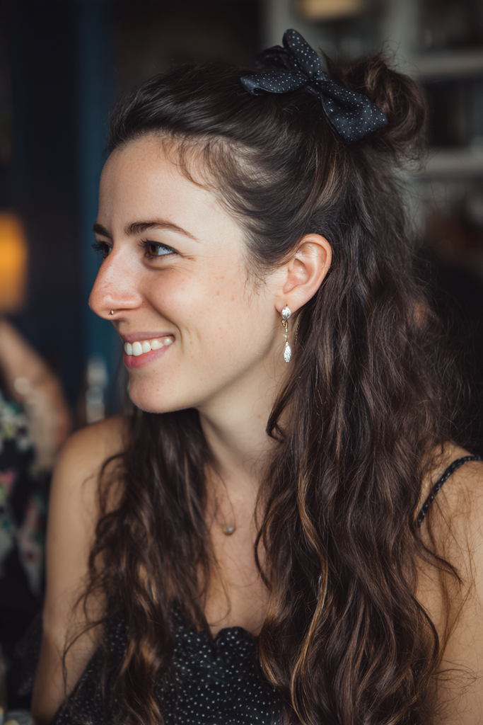A woman with long wavy dark brown hair styled in a half up high bun accessorized with a black polka dot bow.
