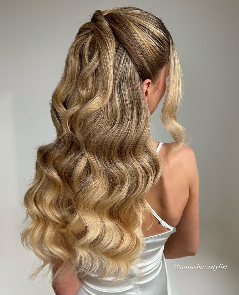 A woman with long dimensional blonde hair styled in glamorous half up waves with face framing pieces.