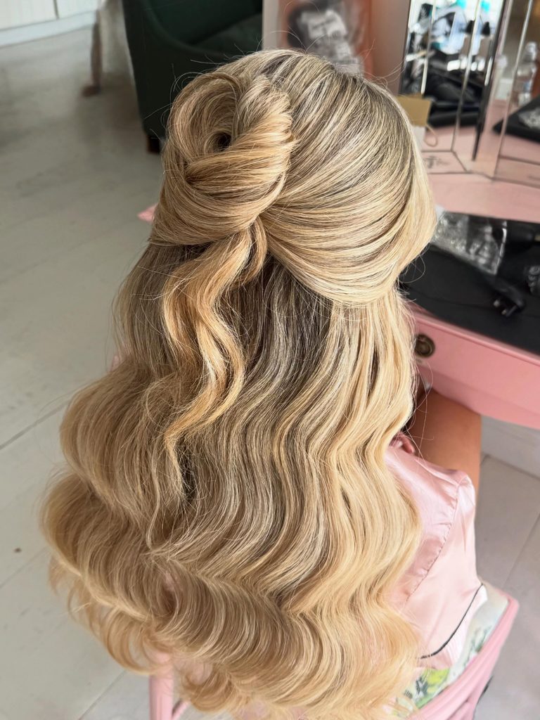 Long blonde hair styled in a half up bun knot with shiny uniform waves falling down the back.